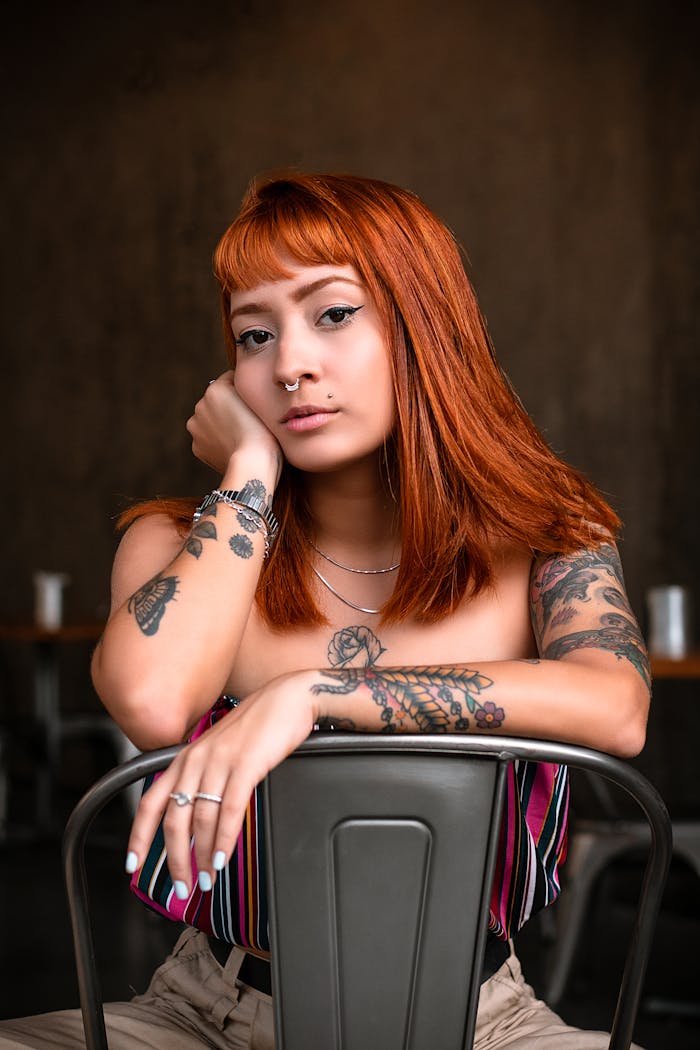 Portrait of a young tattooed woman with red hair posing on a chair indoors.
