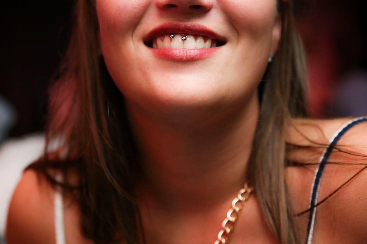 A close-up of a woman smiling, showcasing a unique piercing and a gold chain necklace.