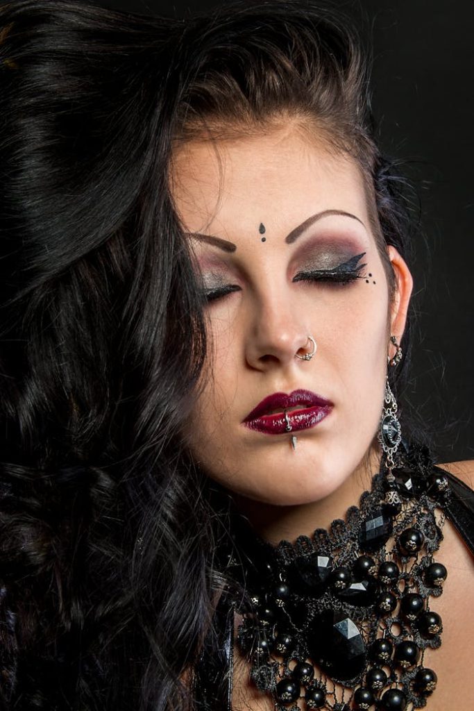 Close-up of an adorned woman with gothic makeup, jewelry, and closed eyes on a dark background.